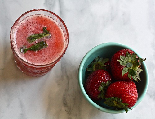 A glass of strawberry cocktail topped with mint leaves and a small bowl of strawberries.