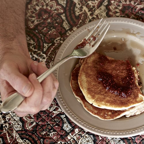 A hand holds a fork over a plate of pancakes.