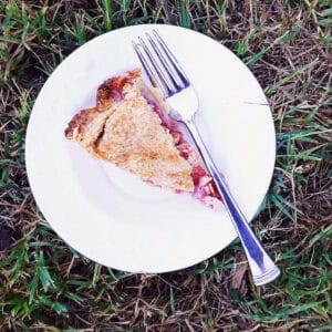 A piece of strawberry rhubarb pie with honey on a white plate with a fork.