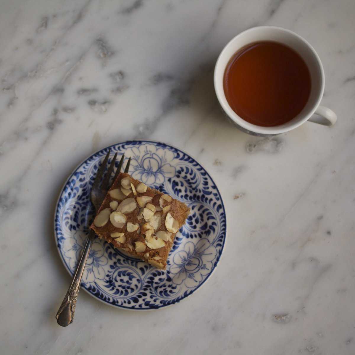 A piece of spelt honey cake on a plate with a fork and a cup of tea next to it.