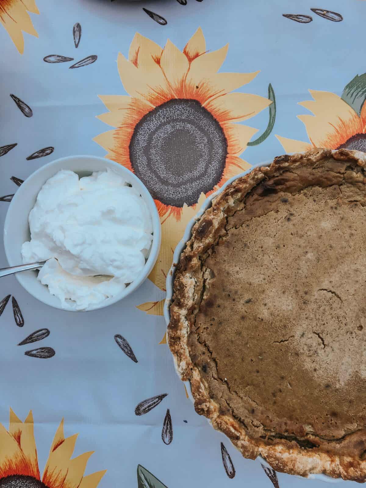 A maple pumpkin pie is seen on a sunflower tablecloth with a bowl of whipped cream to its right.
