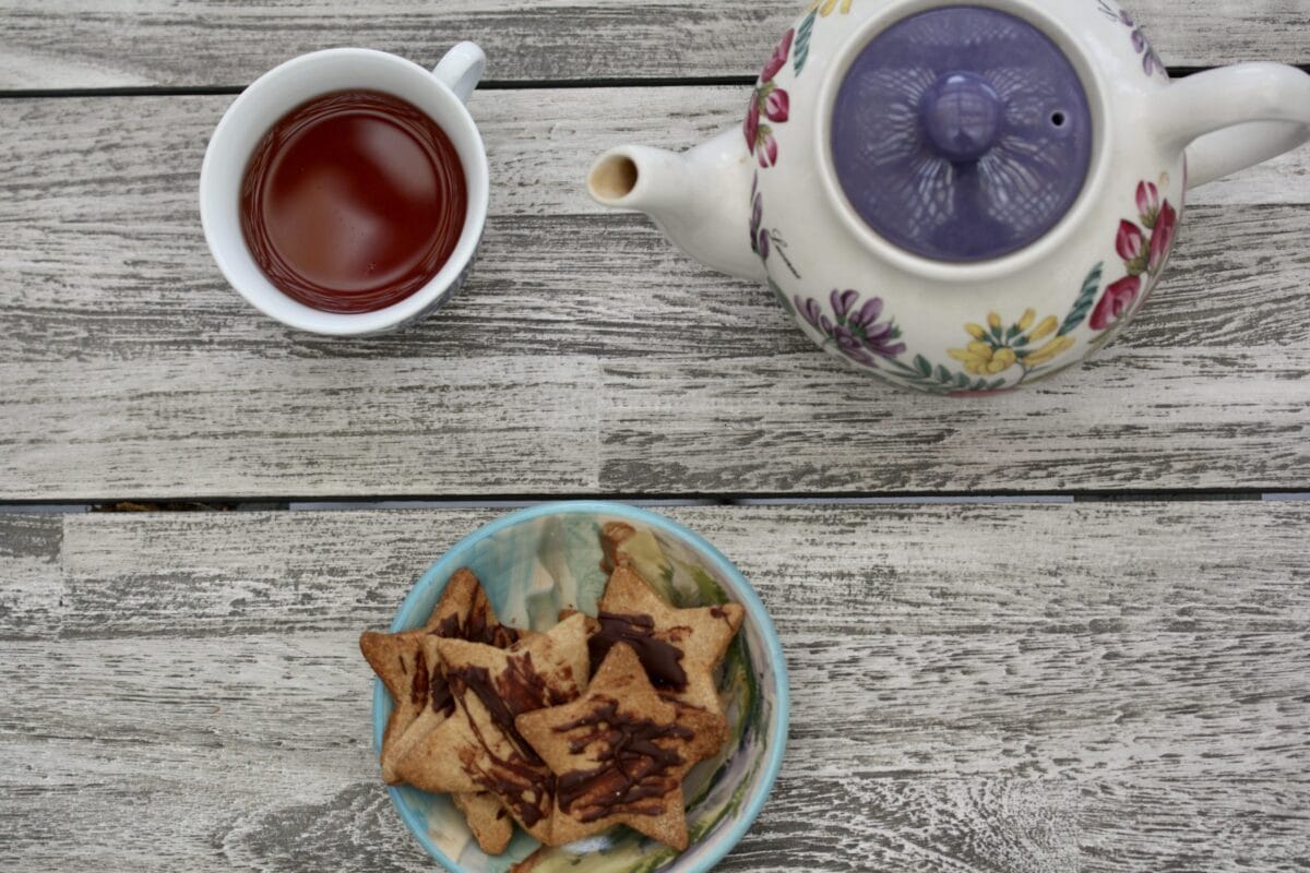 A floral tea pot is shown on a gray table next to a cup of tea with a small dish of shortbread cookie stars below it.