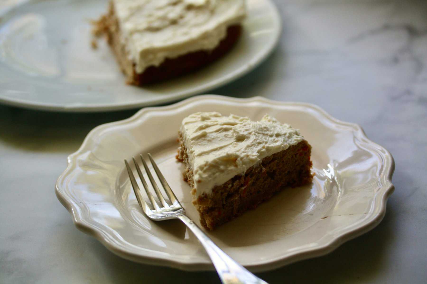 A slice of carrot cake on a plate with a fork with a carrot cake seen in the background.