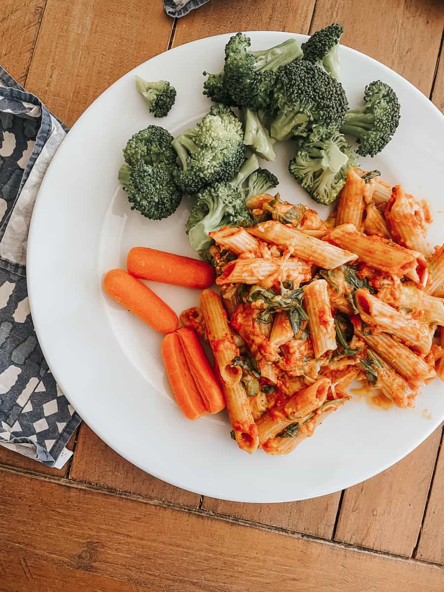 A portion of gluten free pasta bake with broccoli and baby carrots on a white plate.