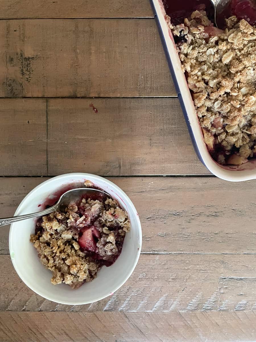 A bowl of gluten free apple blackberry crumble next to a pan of crumble on a wood table.