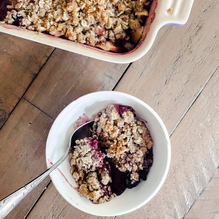 A bowl of apple blackberry crumble next to a pan of crumble on a wood table.