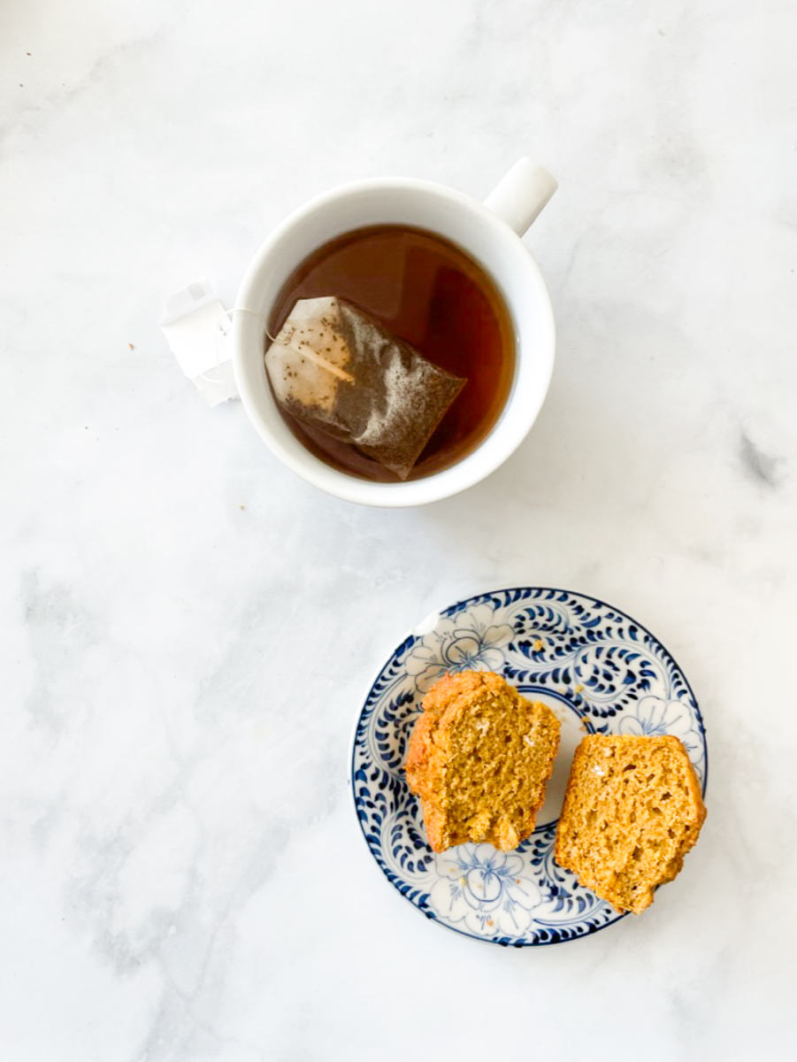 A cup of tea with a tea bag is shown on a white background next to a cup-open gluten-free pumpkin muffin on a blue and white plate.