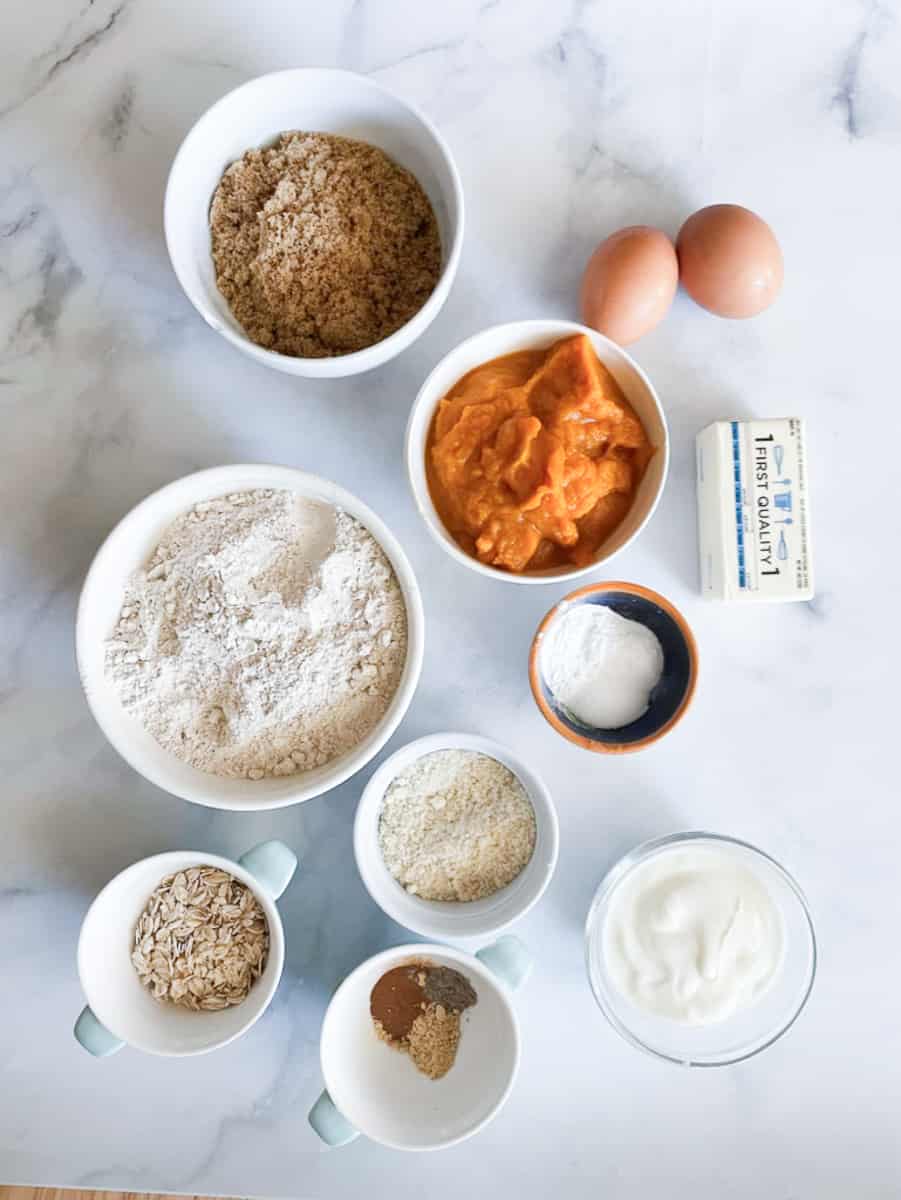 The ingredients for gluten-free pumpkin muffins are shown portioned out: pumpkin puree, butter, eggs, oat flour, oats, almond flour, brown sugar, yogurt, baking soda and baking powder, salt, and spices.