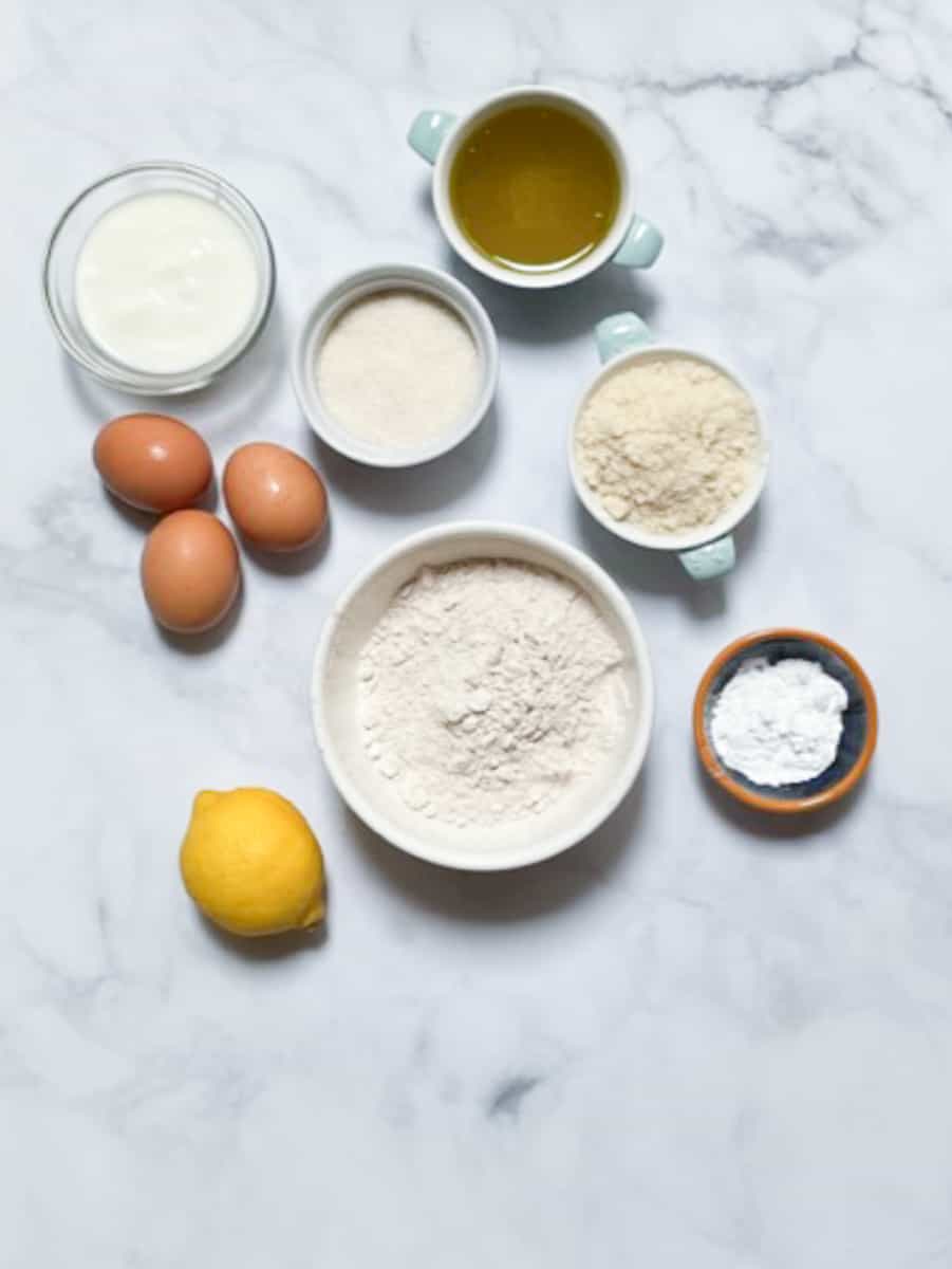 The ingredients for gluten free lemon loaf, including oil, lemons, sugar, oat flour, almond flour, yogurt, and eggs, are shown on a white background.