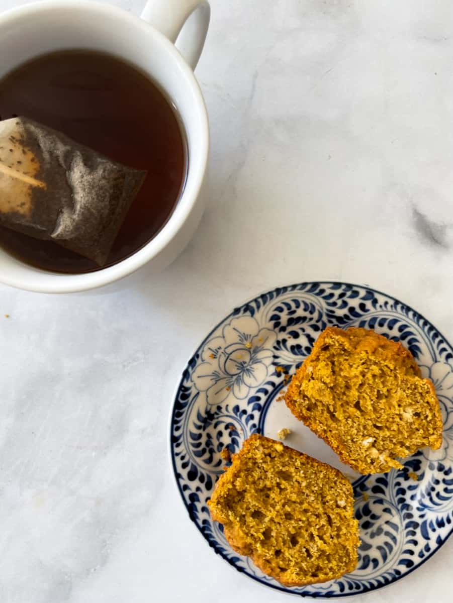 A cup of tea with a tea bag is shown on a white background next to a cup-open gluten-free pumpkin muffin on a blue and white plate.