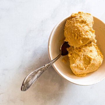 A bowl of scoops of pumpkin ice cream with a spoon is shown on a white background.