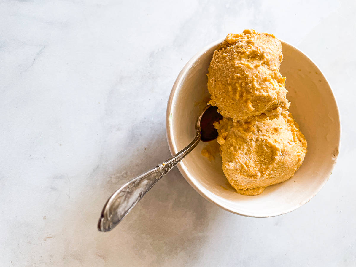 A bowl of scoops of pumpkin ice cream with a spoon is shown on a white background.