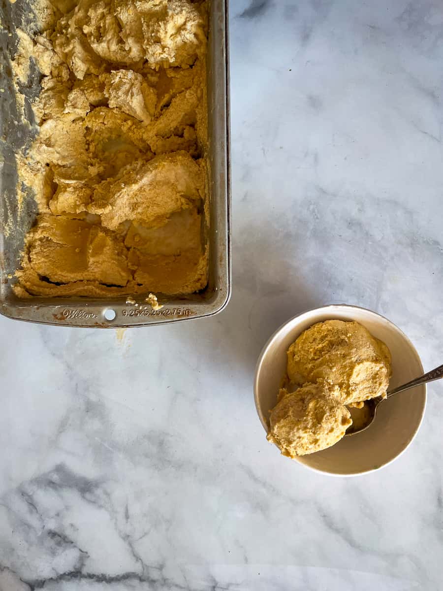 A pan of pumpkin ice cream is shown on a white background with a bowl of pumpkin ice cream next to it.