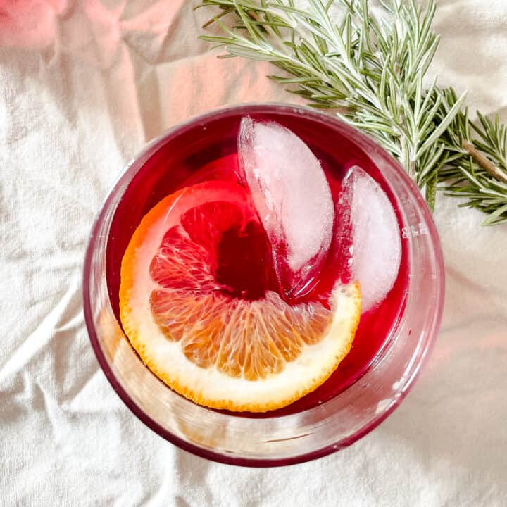 A cranberry margarita in a glass with an orange slice and garnished with rosemary.