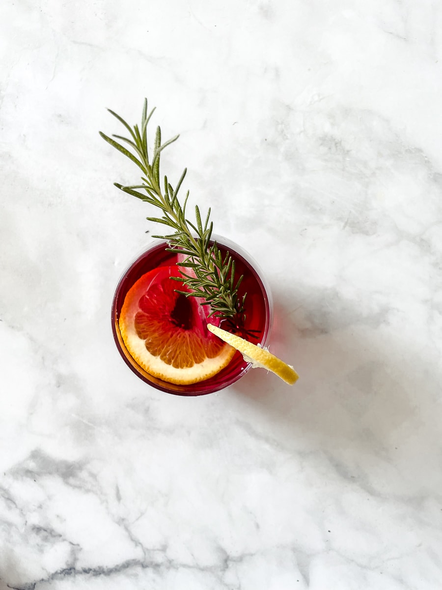 A cranberry margarita in a glass with an orange slice and lemon and garnished with rosemary.