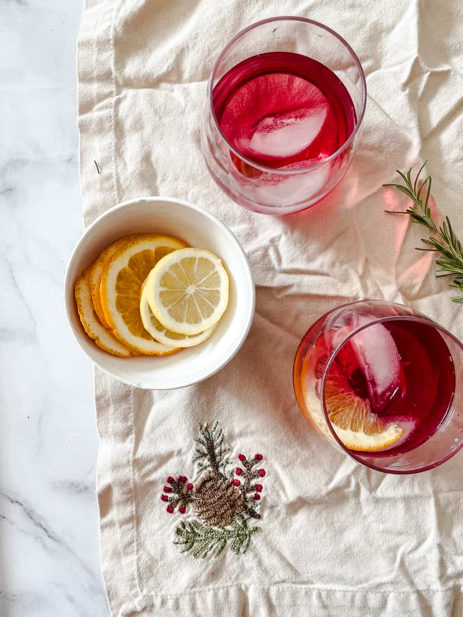 Glasses of cranberry margarita on a festive napkin next to a bowl of orange and lemon slices.