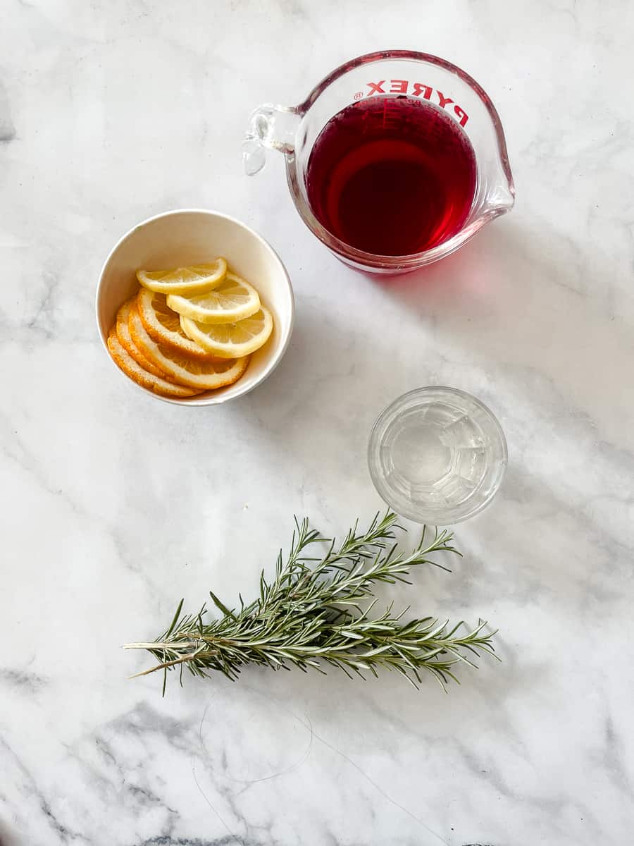 Ingredients needed to make a cranberry margarita are shown: tequila, cranberry juice, lemon and orange slices, fresh rosemary.
