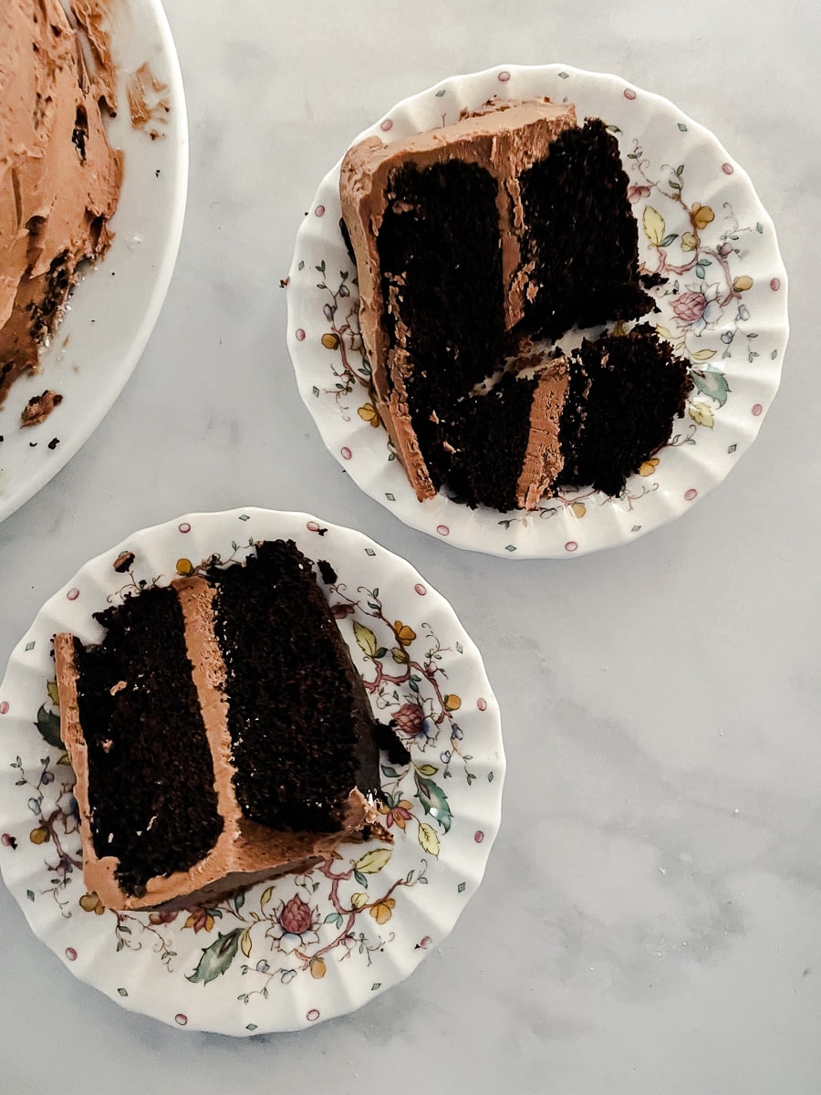 Two slices of gluten free chocolate layer cake on floral plates with the cake visible to their left.