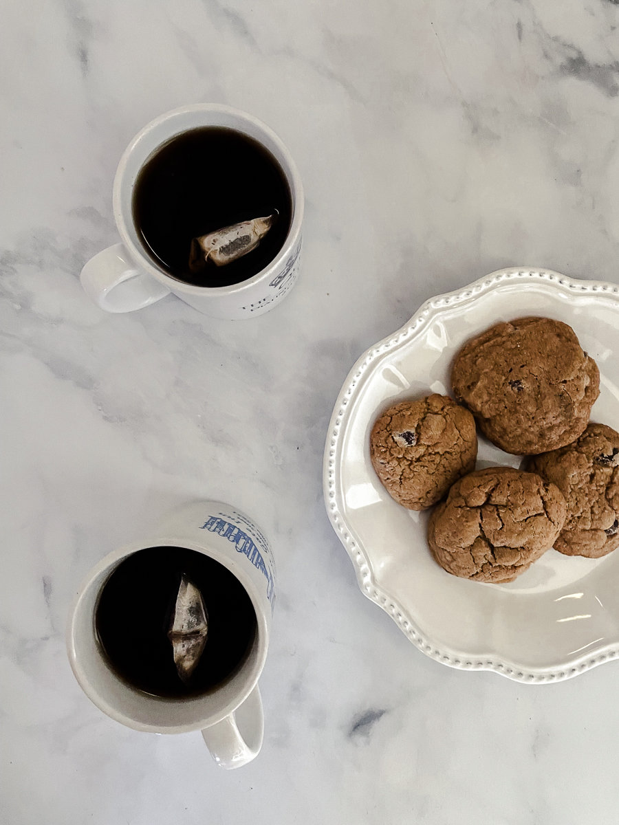 Two mugs of tea with tea bags in them next to a plate of whole wheat chocolate chip cookies.