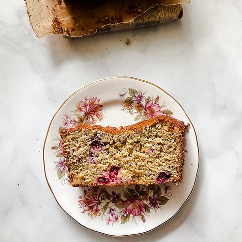 A slice of cranberry orange bread on a floral plate with the loaf nearby.