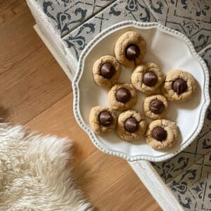 A plate of gluten free peanut butter blossoms site on a fireplace above a wood floor showing a white wool rug.