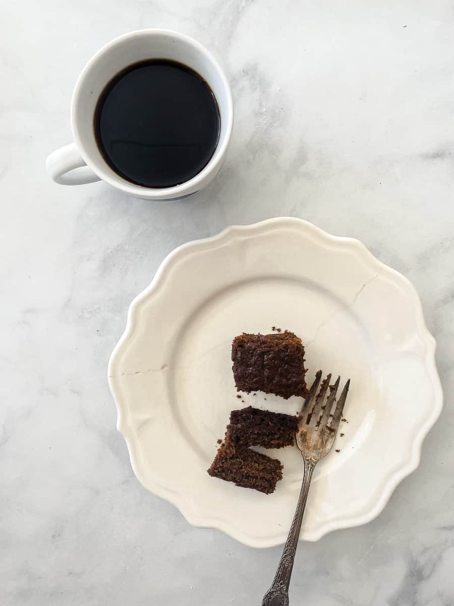 A cut piece of gluten free gingerbread on a white plate with a black cup of coffee next to it.