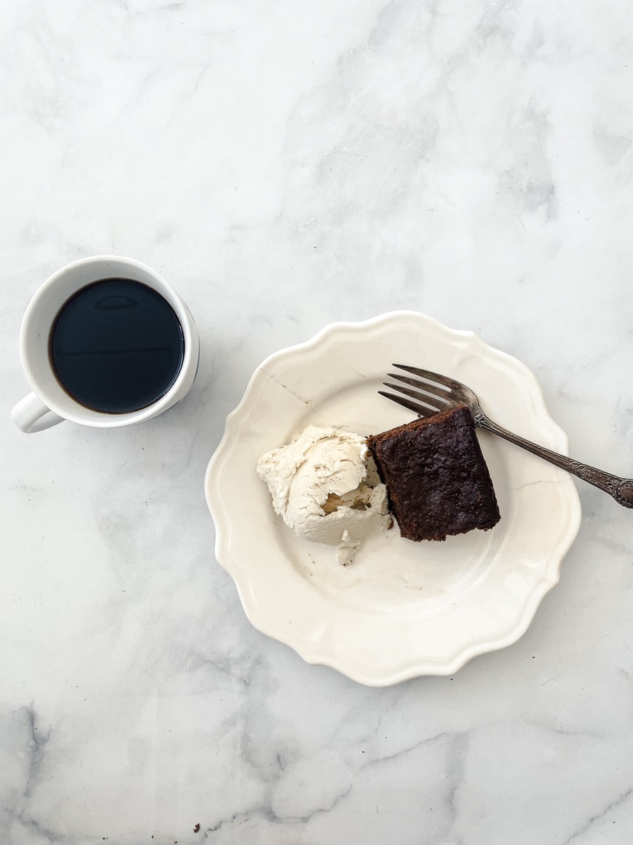A cup of coffee on a white background to the left of a white plate containing a slice of gluten free gingerbread and ice cream.