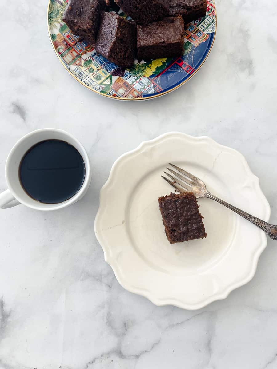 Half of a plate of stacked gluten free gingerbread pieces are shown with a cup of coffee and a square of gingerbread on a white plate with a fork.