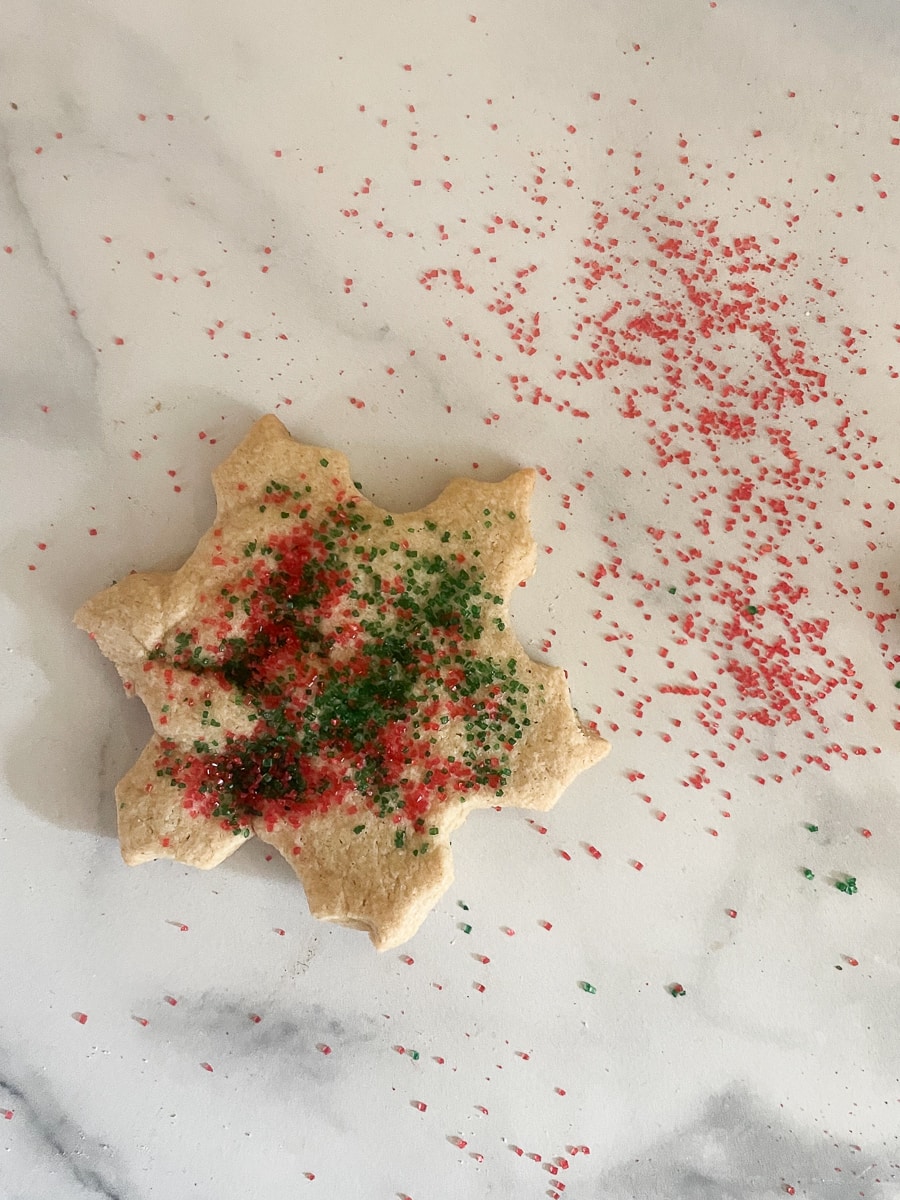 An oat flour sugar cookie in the shape of a snowflake on a white background with red sprinkles scattered around.