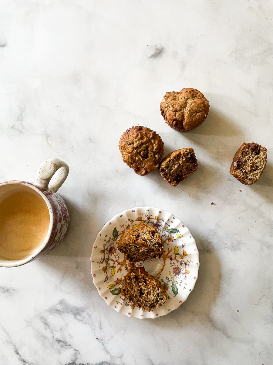 A latte, a plate holding a cut open muffin, and more oat flour banana muffins around them.