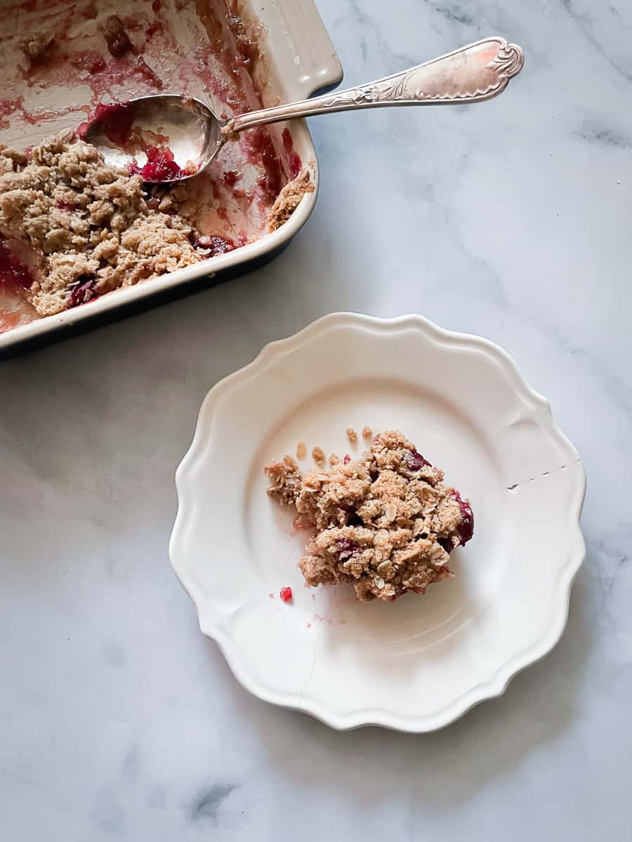 A portion of cranberry apple crisp on a plate next to a pan of crisp.