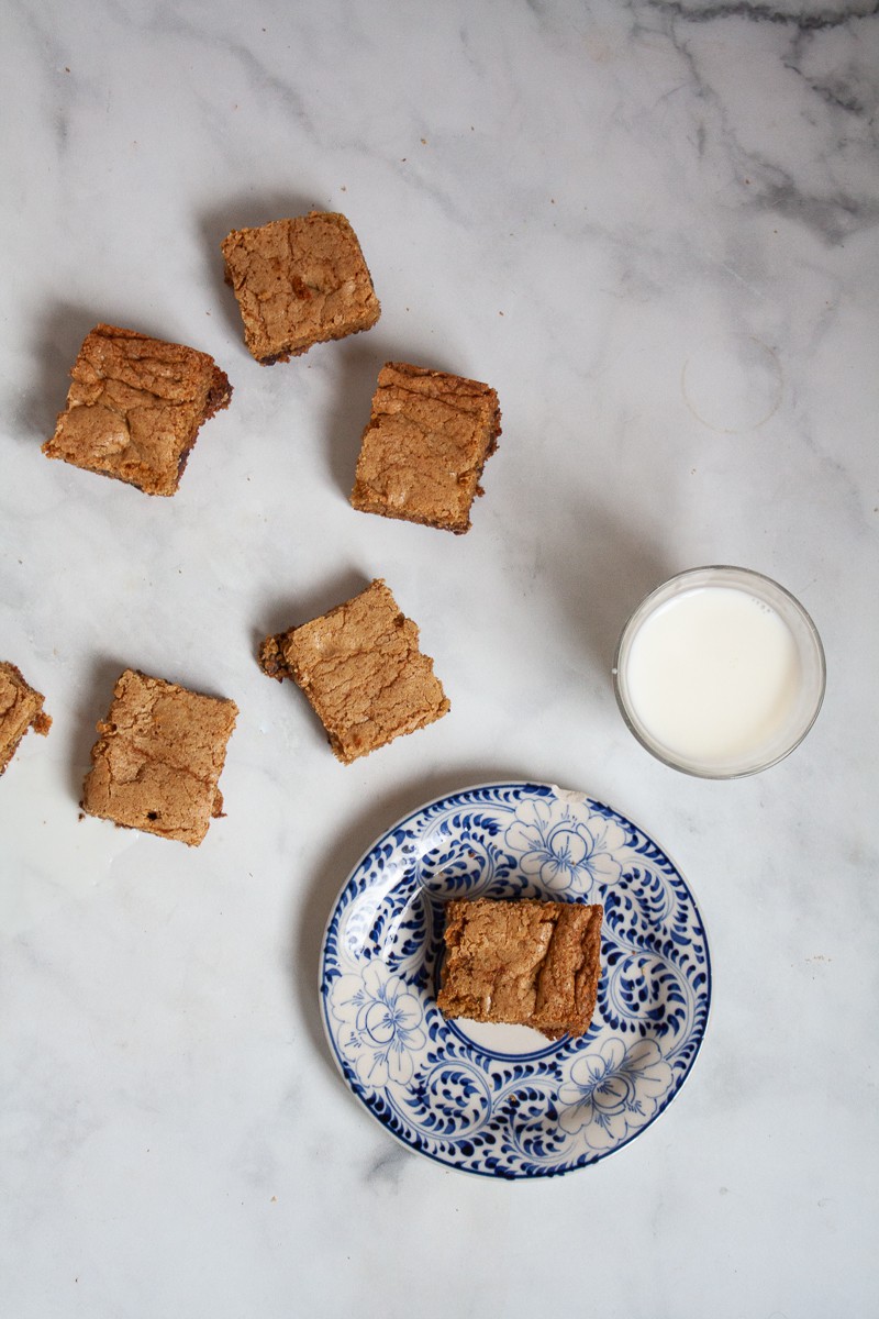 Chocolate chip cookie bars on a white counter with a glass of milk and a plate with one bar.