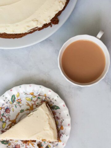 A piece of gluten free carrot cake on a plate with a cup of tea and the cake shown behind it.