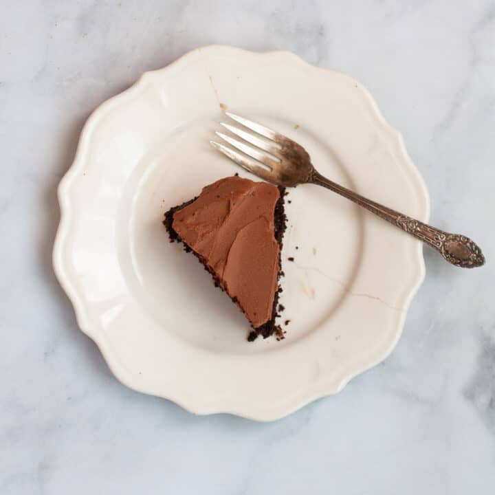 A white plate holds a slice of oat flour chocolate cake with a fork alongside.