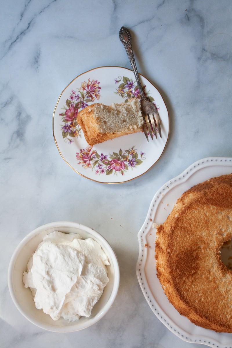 Half of an angel food cake is seen next to a bowl of whipped cream and a slice of cake on a plate.