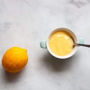 A lemon rests to the left of a dish of honey lemon curd with a spoon in it.