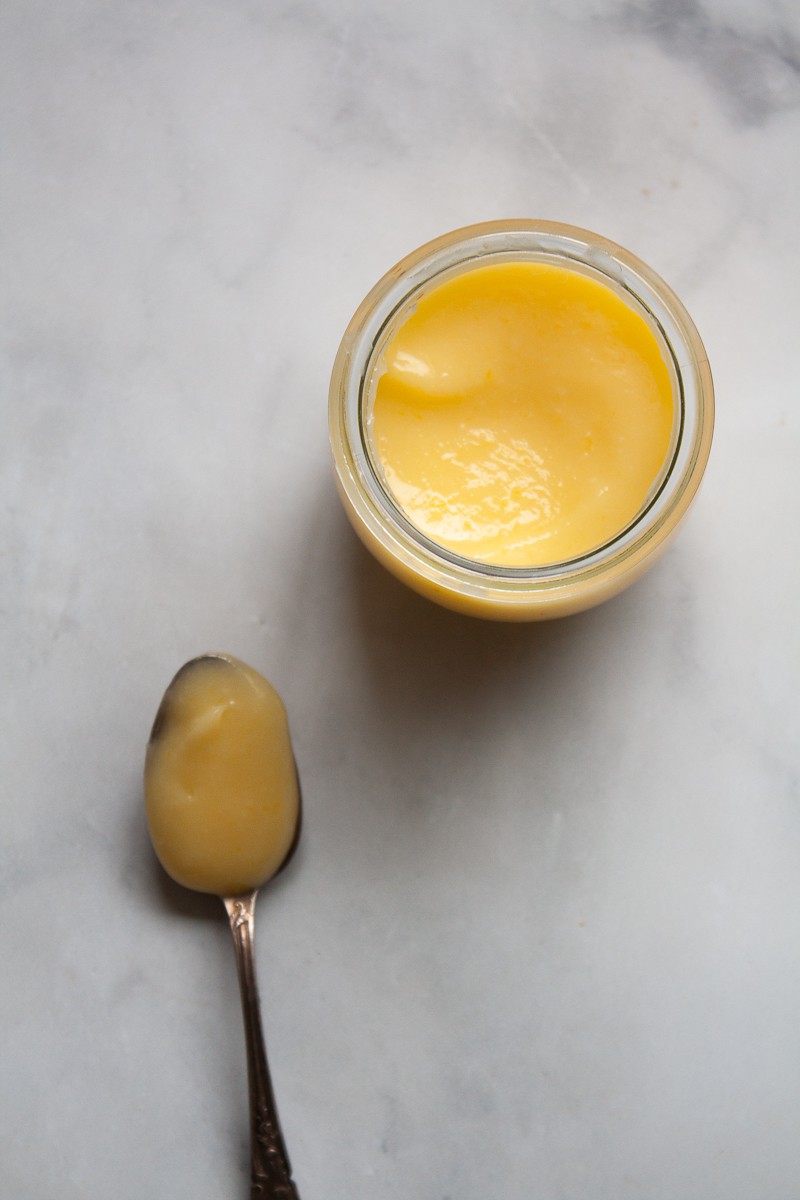 A spoon of lemon curd next to a jar of lemon curd.