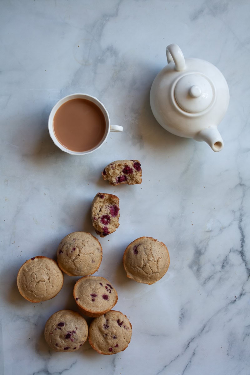 A cup of tea, a white tea pot, and oatmeal raspberry muffins.