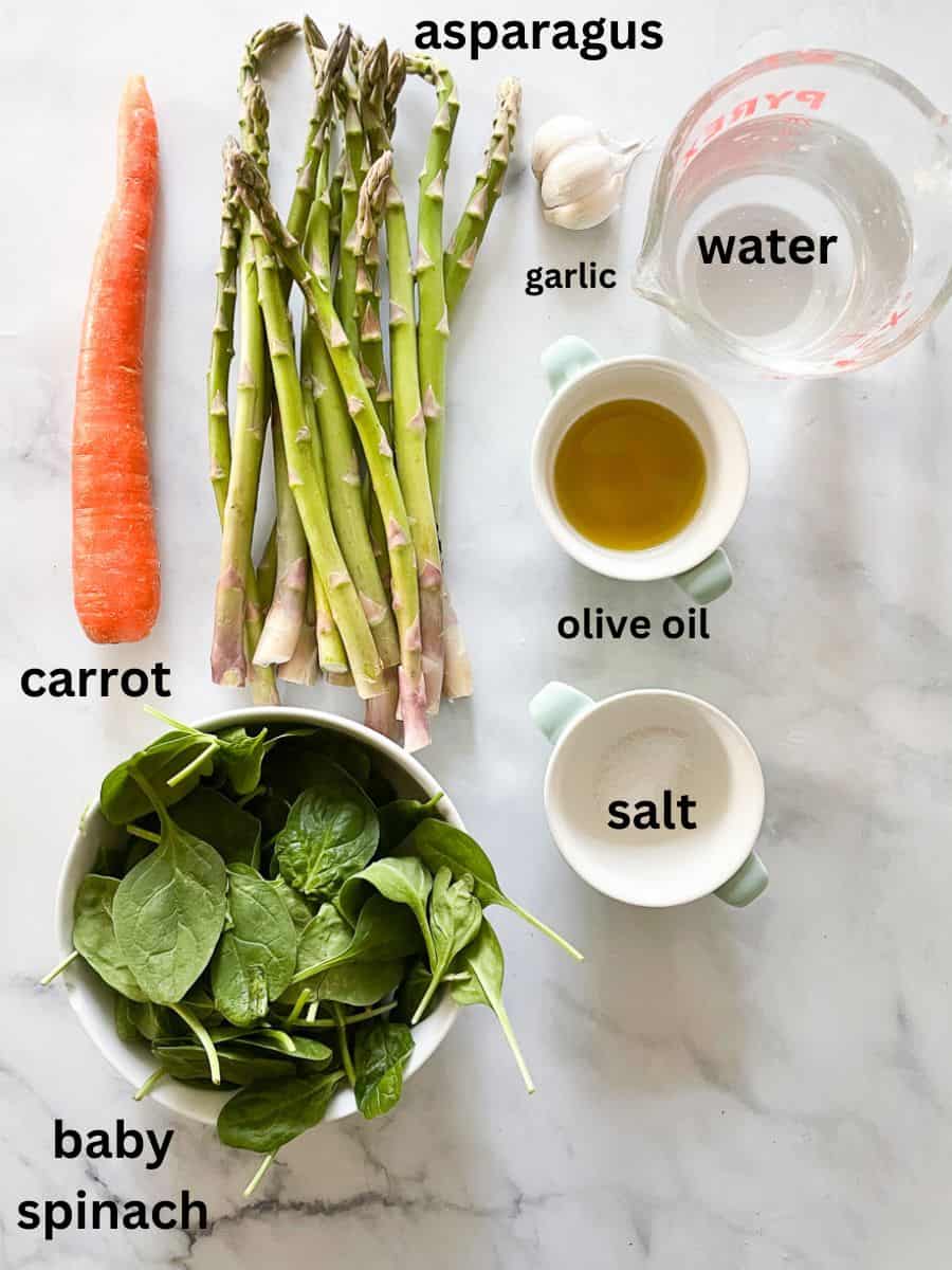 Ingredients for vegan asparagus soup are shown labelled and portioned out: asparagus, carrot, salt, olive oil, water, spinach, and garlic.