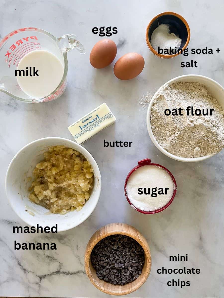 Ingredients for gluten free banana chocolate chip cake are labelled and portioned out: oat flour, baking soda, milk, sugar, mashed banana, eggs, mini chocolate chips, butter.