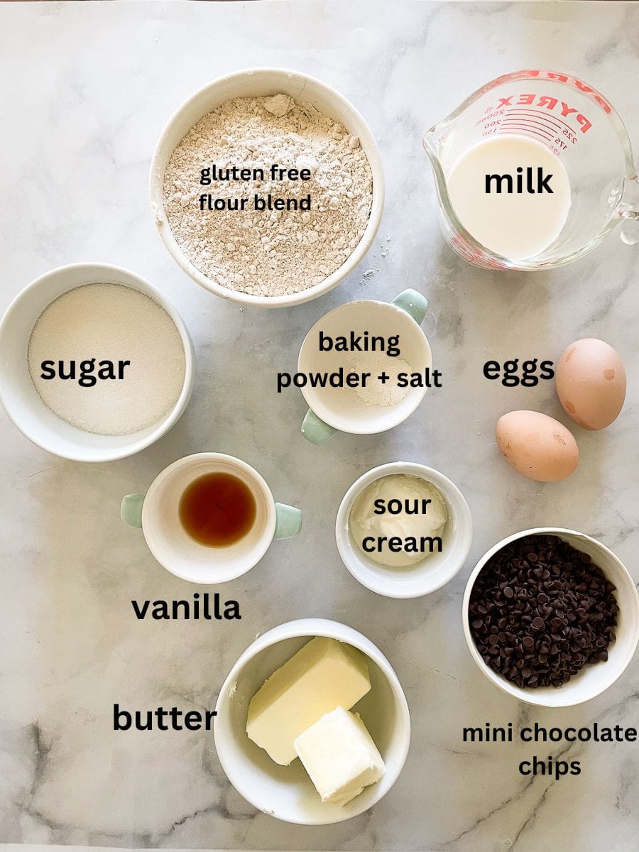 Ingredients for gluten free chocolate chip loaf cake are portioned out and labelled: gluten free flour blend, baking powder, salt, vanilla, eggs, sour cream, milk, mini chocolate chips, butter.