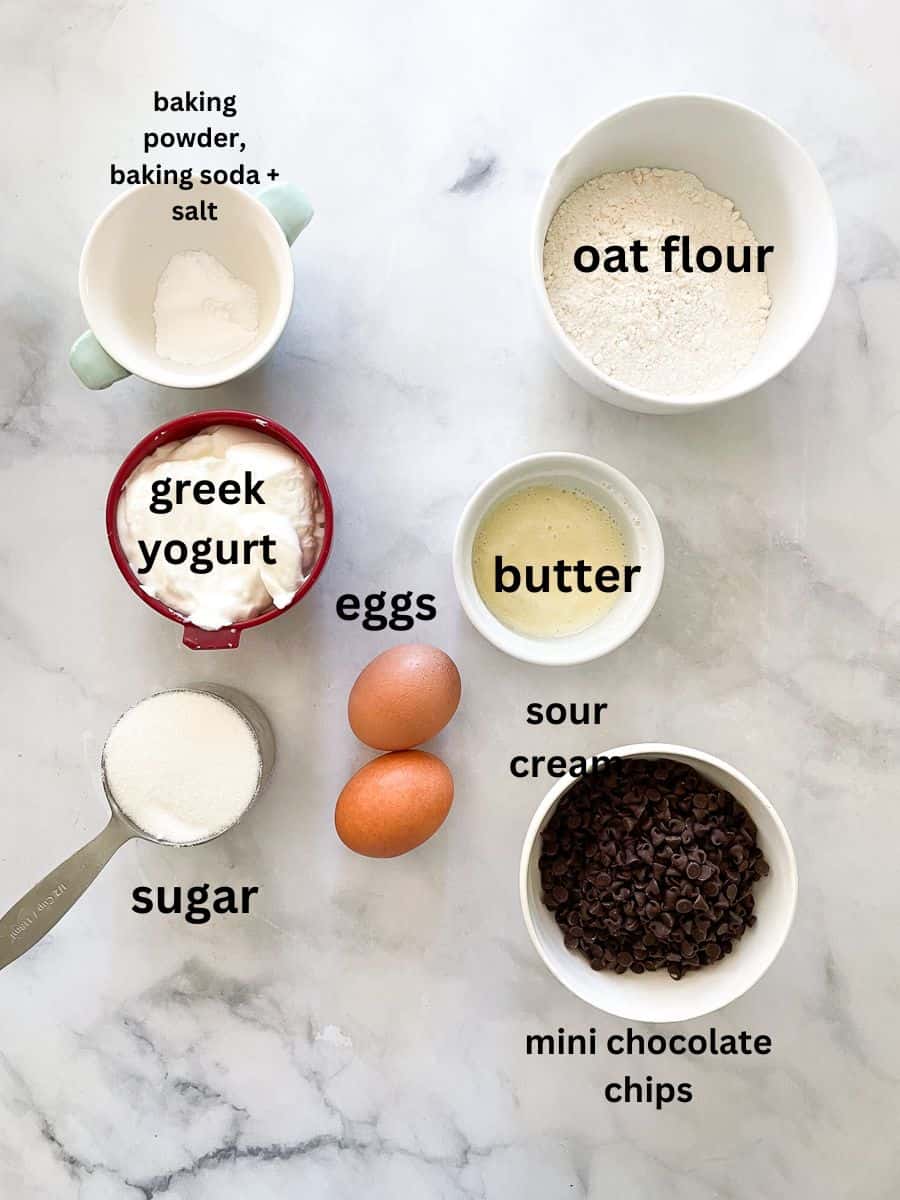 The ingredients for gluten free mini chocolate chips muffins are labelled and portioned out: mini choco chips, oat flour, greek yogurt, eggs, sugar, butter, baking powder, baking soda, salt.