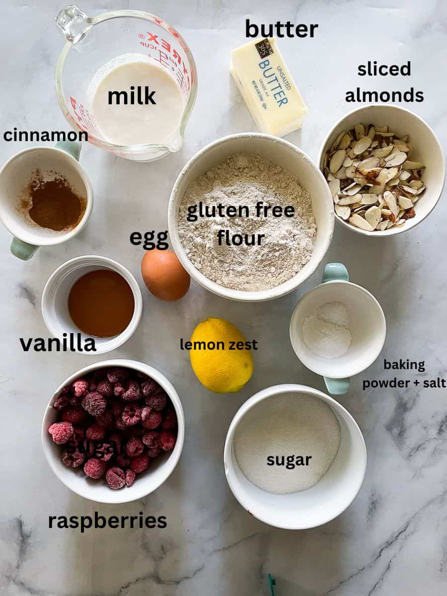 The ingredients for gluten free raspberry almond cake are portioned out and labelled: lemon, milk, cinnamon, gluten free flour, almonds, vanilla, raspberries, sugar, baking powder, salt.