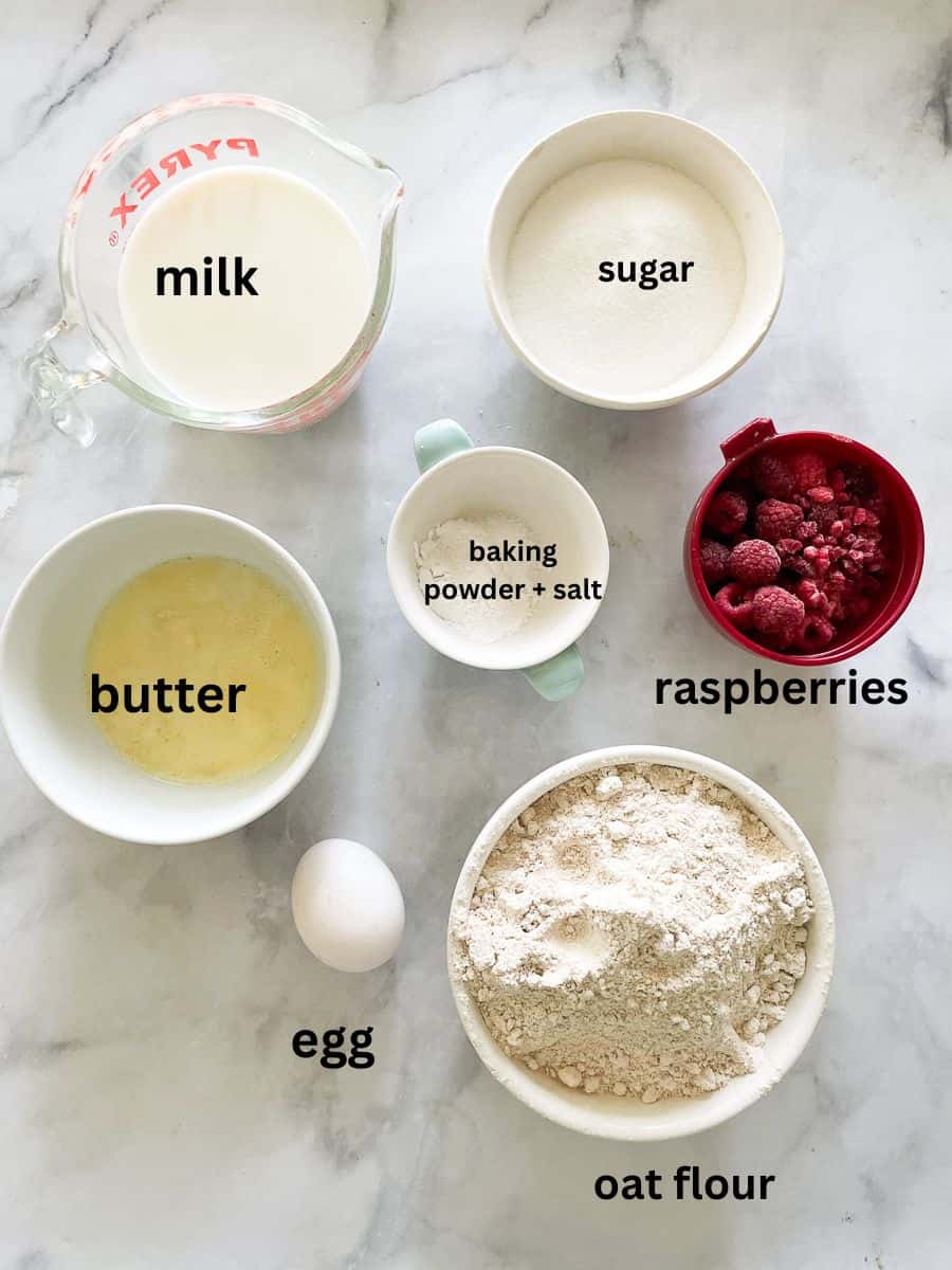 The ingredients for oatmeal raspberry muffins are shown labelled and portioned out: oat flour, raspberries, sugar, butter, egg, baking powder, salt, milk.