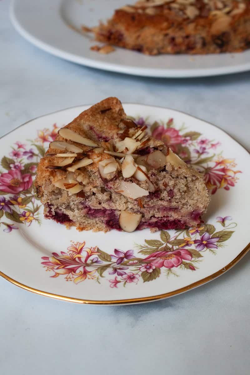A piece of raspberry almond cake on a plate with the cake in the background.