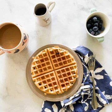Gluten free waffles for two on a plate with coffee, a pitcher of maple syrup, and a bowl of blueberries.