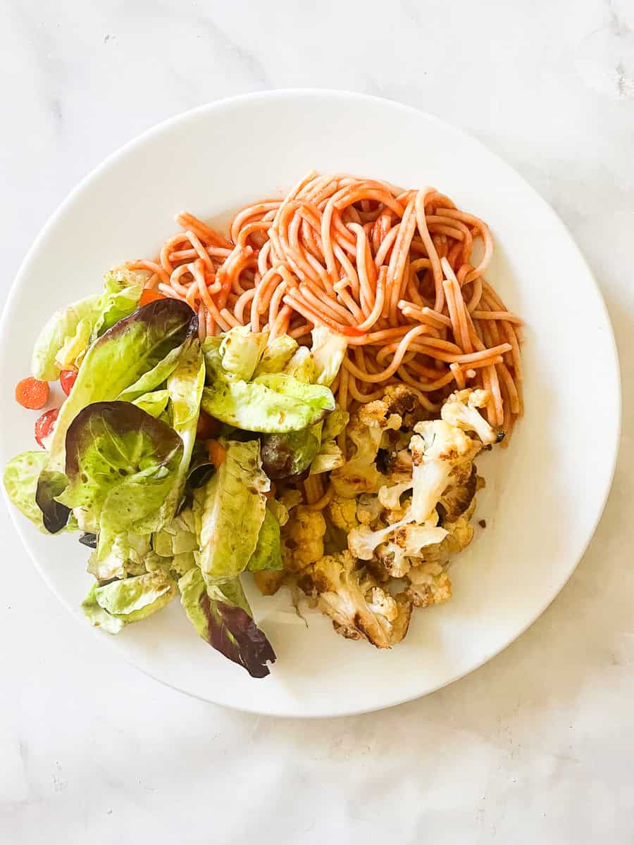 A plate holds spaghetti with red sauce, salad, and roasted cauliflower.