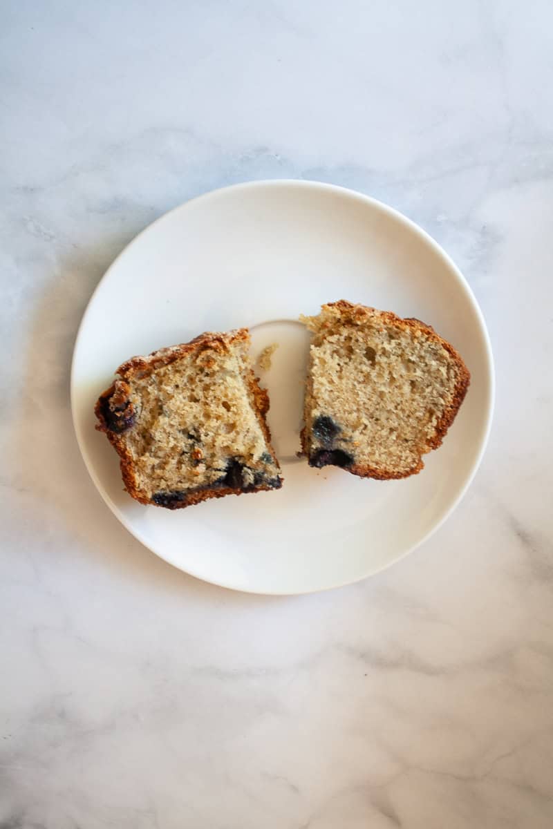 A piece of gluten free blueberry bread is cut in a half on a white plate.