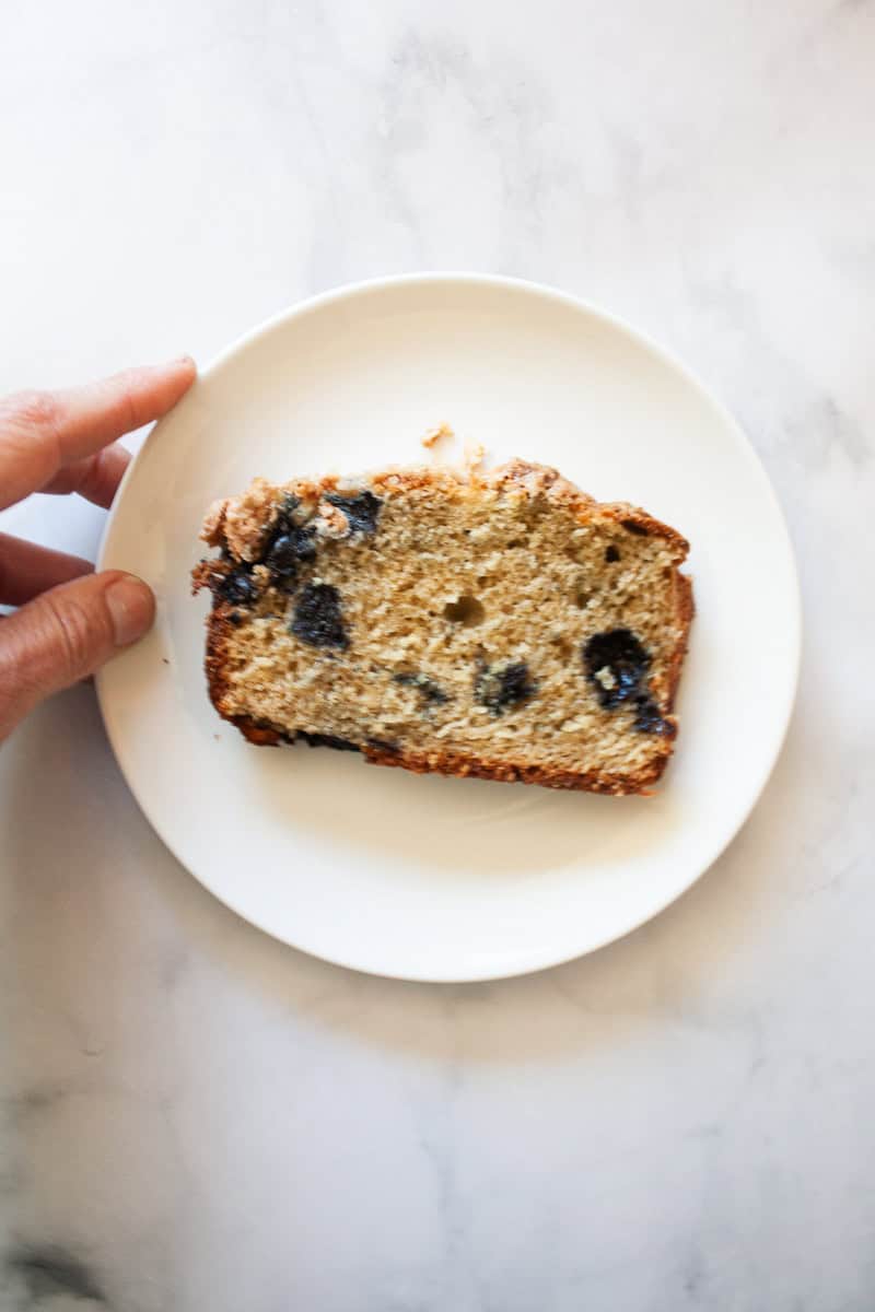 A slice of gluten free blueberry bread is served on a white plate with a hand adjusting the plate.