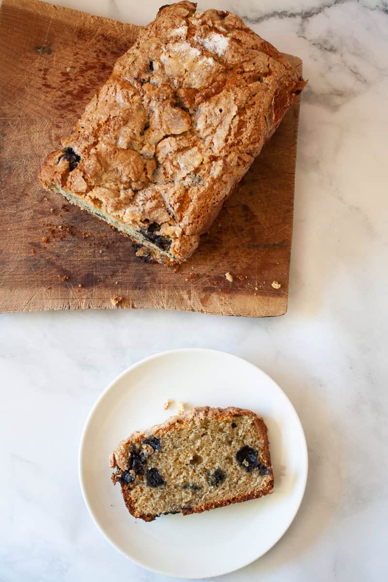 A slice of gluten free blueberry bread on a plate next to the loaf.